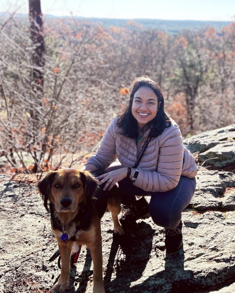 Asrah Natalie Mohammed crouching on a rock outcrop beside her dog Boone, with autumn woods and an open valley behind them